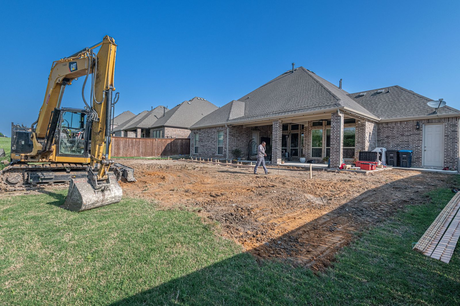 Excavator and string lines laying out a new backyard patio behind a Texas brick house